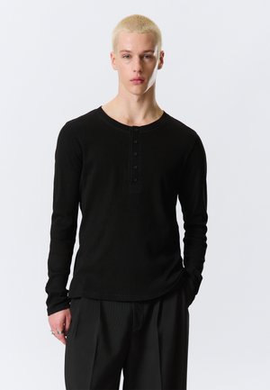Jeune personne aux cheveux blonds courts portant une chemise henley noire à manches longues et un pantalon noir, debout devant un fond clair uni.