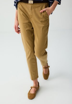 Personne portant un pantalon fuselé beige, une ceinture marron, des chaussures Mary Jane marron et un haut rayé bleu, une main dans la poche en marchant.