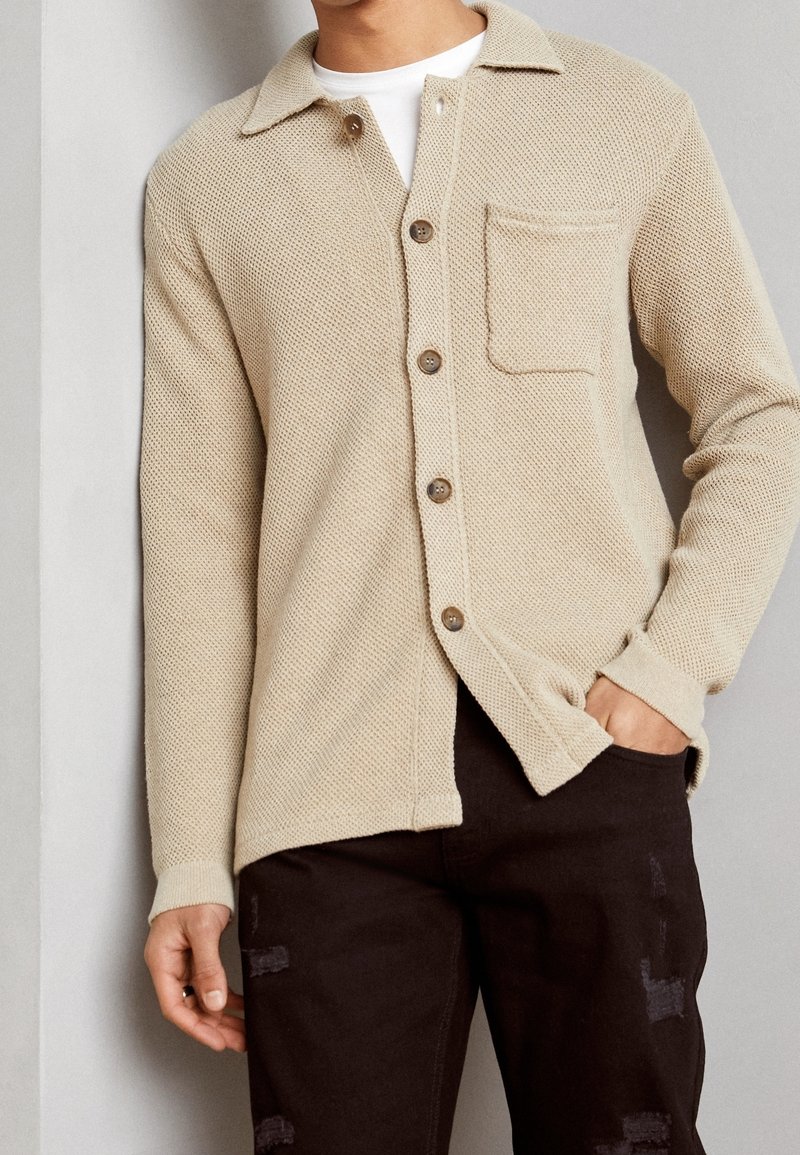 Cárdigan de punto beige con cuello, cinco botones y un bolsillo en el pecho. Puños acanalados y un acabado texturizado, combinado con pantalones oscuros.