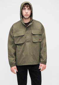 Μπουφάν με κουκούλα σε ελιά πράσινο χρώμα, κατασκευασμένο από ελαφρύ υλικό, με δύο τσέπες στο στήθος και πλευρικές ανοίγματα. Διαθέτει ραφές διακοσμητικές στα μανίκια.