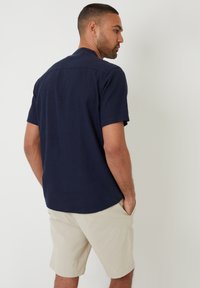 Korte mouwen, donkerblauwe shirt met een rechte zoom en subtiele textuur, gecombineerd met lichtbeige shorts, handen in de zakken.