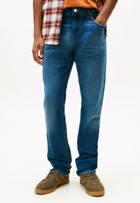 Jeans de mezclilla azul oscuro con corte recto, que presentan zonas deslavadas para dar textura. Combinados con zapatillas marrones y una camisa de franela a cuadros.