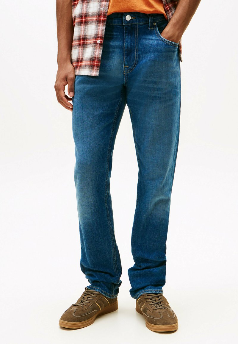 Jeans de mezclilla azul oscuro con corte recto, que presentan zonas deslavadas para dar textura. Combinados con zapatillas marrones y una camisa de franela a cuadros.