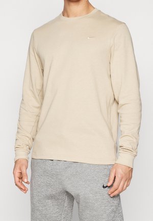 Homme portant un t-shirt beige manches longues Nike et un pantalon de jogging gris Nike, debout avec les mains détendues le long du corps sur un fond uni.