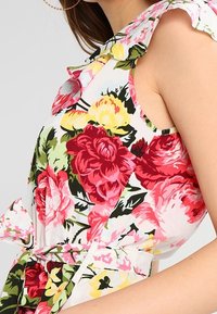 Floral print dress in white fabric. Features red and yellow roses, ruffled shoulder straps, and a tied waist for a fitted silhouette.