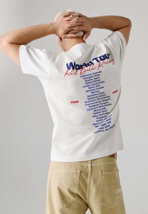 Mann mit weißen Haaren, der ein weißes Red Bull Racing World Tour T-Shirt mit den für 2026 aufgeführten Rennorten trägt und mit den Händen am Kopf posiert.