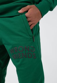 Groene sweatpants met een gladde textuur, met een zwarte grafische tekst "WRONG FRIENDS" op het linkerpoot en een zippocket aan de zijkant.