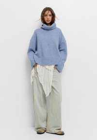 Világoskék kötött pulóver, magas garbónyakkal és oversized ujjal, világos krémszínű anyag felett. Világoskék farmernadrággal és barna lábbelivel párosítva.