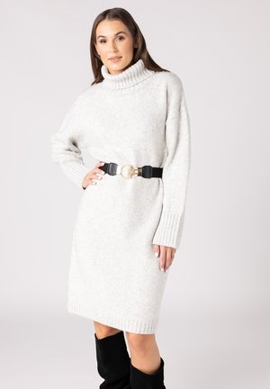 Lichtgrijze oversized trui jurk met een funnelneck, geribbelde manchetten en een zwarte riem met een gouden gesp, gedragen met zwarte kniehooglaarzen.