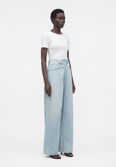 Nő fehér rövid ujjú ingben és világoskék, széles szárú farmerral, egyszerű fehér háttér előtt áll.