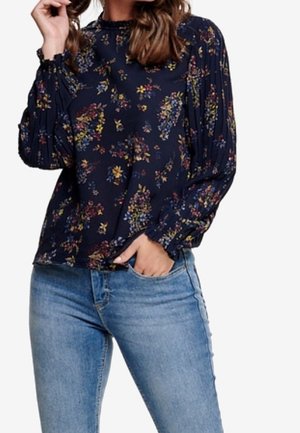 Woman wearing a navy floral long-sleeve blouse with ruffled cuffs and light blue jeans, one hand in pocket and the other near her face.