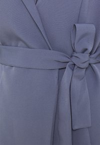 Měkká, levandulová látka s hladkým povrchem. Obsahuje zavázaný opasek v pase pro uzavření a otevřený klopu design.