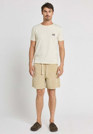 Homme debout, portant un short beige, un t-shirt crème avec un petit logo animal sur la poitrine et des chaussures marron foncé sans lacets, sur fond uni.