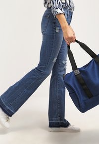 Modré denimové zvonové džíny s odřeným detailem, v kombinaci s texturovanou modrou taškou s černými popruhy a bílými teniskami.