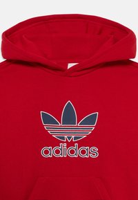 Raudona medvilninė džemperis su gobtuvu, puošta mėlynu siuvinėtu Adidas logotipu su trefoil simboliu ir trimis horizontaliosiomis juostomis. Savyje turi priekinę kišenę.