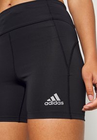 Shorts de sport noirs à texture lisse, avec des coutures contrastantes et un logo Adidas argenté sur la partie inférieure de la jambe gauche.