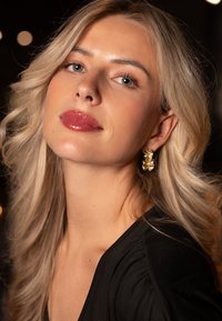 Guldteksturerede øreringe med et skulpturelt design, båret af en kvinde med langt, bølget blond hår, mod en sløret mørk baggrund.