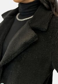 Black fleece coat with a textured surface, featuring a notched lapel design and visible stitching. Layered with a black turtleneck underneath.