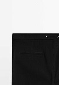 Gros plan de la ceinture d’un pantalon noir avec une poche fendue à l’arrière et un petit ruban en dentelle passé à travers des œillets métalliques sur le côté droit.