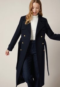 Granatowy płaszcz trencz wykonany z mieszanki wełny, z podwójnym zapięciem, złotymi guzikami, szerokim kołnierzem oraz kieszeniami po bokach. W zestawie z białym golfem i ciemnymi dżinsami.