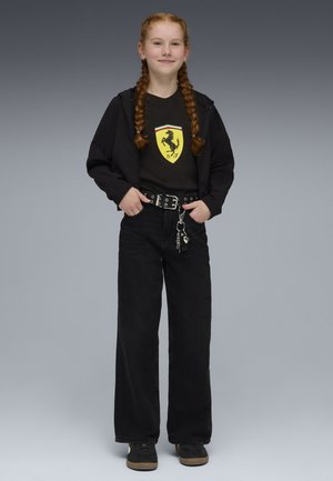 Dziewczyna z czerwonym, zaplecionym warkoczem, ubrana w czarną koszulkę z logo Ferrari, czarną kurtkę, szerokie czarne spodnie oraz czarne buty z gumową podeszwą.