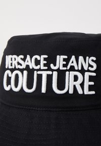 Melna ķiveres cepure ar izšūtu balto tekstu "VERSACE JEANS COUTURE", ar teksturētu audumu un strukturētu malas daļu.