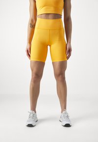 Žuti atletski crop top i visoki biciklistički šorts izrađeni od rastezljivog materijala. Model nosi bijele tenisice za trčanje s sivim i plavim detaljima.