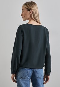 Dunkelgrüne Bluse mit langen Ärmeln und rundem Halsausschnitt, glatte Textur. Kombiniert mit hellblauen Denim-Jeans. Schlichtes Design mit knöpfbaren Manschetten.