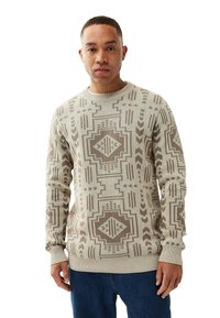 Junger Mann in einem beigefarbenen und braun gemusterten Strickpullover und blauen Jeans, der vor einem weißen Hintergrund steht.