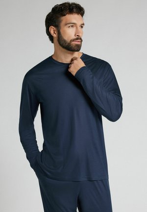 Hombre con barba vistiendo camisa de manga larga azul marino y pantalones a juego, una mano cerca del cuello, mirando hacia un lado contra un fondo liso.