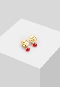 Los pendientes en tono dorado cuentan con discos circulares texturizados y acentos de cuentas rojas colgantes. El diseño es moderno y llamativo, con colores en contraste.