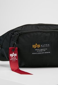 Svart nylon midjeväska med dragkedjefack, en röd tagg med logotyp och en vävd etikett på framsidan. Texturerad yta.