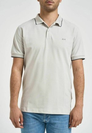 Hombre con polo de manga corta gris claro con pequeño logo en el pecho izquierdo y vaqueros azules, de pie frente a un fondo blanco liso.