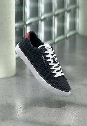 Baskets en mesh noir avec semelle et lacets blancs, détail rouge au talon, et marque Tommy Hilfiger, flottant au-dessus d'un sol poli.