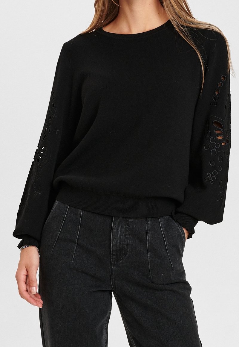Femme portant un pull noir avec des motifs floraux découpés sur les manches bouffantes et un jean foncé taille haute, main dans la poche.