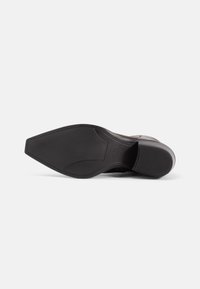 Semelle d’une chaussure formelle noire à bout pointu, semelle extérieure en caoutchouc texturé et petit talon empilé sur fond blanc uni.