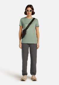 Camiseta verde menta con un estampado gráfico, bolso bandolera negro, pantalones de chándal grises y zapatillas beige. Gafas de sol blancas redondeadas completan el look.