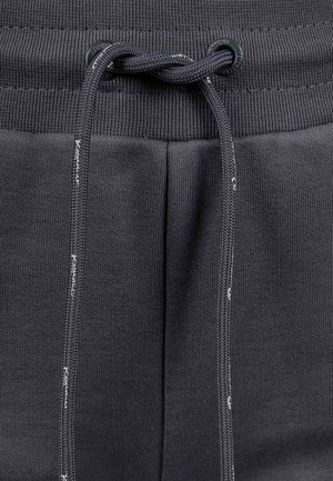 Un pantalon de survêtement gris foncé présente une taille côtelée avec un cordon de serrage, des coutures contrastantes et une texture lisse.