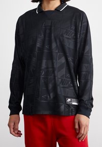 En person som bär en svart långärmad Nike Air-tröja med mönstrade logotyper och röda byxor, visad från halsen till höfterna, med händerna avslappnat vid sidorna.