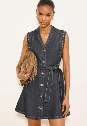 Kvinde iført mørk denimkjole uden ærmer med knapper foran og bælte i taljen, der holder en tan farvet ruskind clutch med frynser.