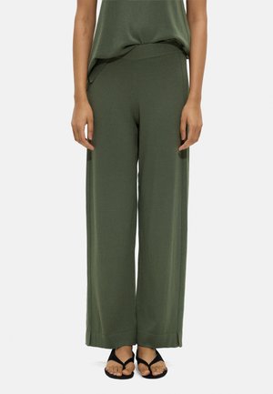 Kobieta w oliwkowych spodniach o szerokich nogawkach, w połączeniu z dopasowanym topem i czarnymi sandałami typu japonek, stojąca na jednolitym tle.