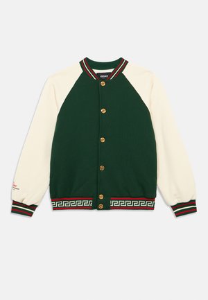 Jachetă varsity verde și crem, cu nasturi aurii, guler și mâneci cu tiv striat, și un model cheie grecească de-a lungul tivului în roșu și verde.