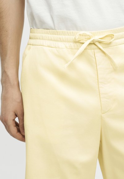 Κοντινό πλάνο ενός ατόμου που φοράει ανοιχτοκίτρινο παντελόνι με κορδόνι και λευκό πουκάμισο, εστιάζοντας στη μέση και το χέρι στο πλάι.