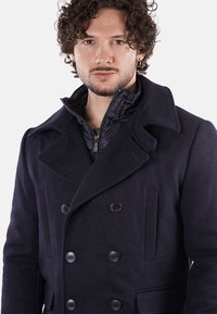 Cappotto peacoat doppiopetto navy con grandi bottoni, colletto a rever e tasche laterali. Presenta una giacca interna con zip per maggiore calore.