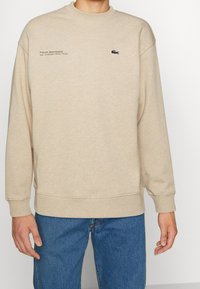 Sudadera beis hecha de una tela suave, con cuello redondo, hombros caídos y el logo de Lacoste bordado en el pecho junto con texto.