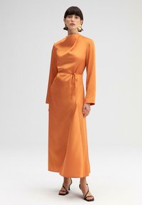 Langdurige oranje satijnen jurk met een hoge halslijn, een tailleriem en een zijsplit, gecombineerd met zwarte sandalen met hak en banden.