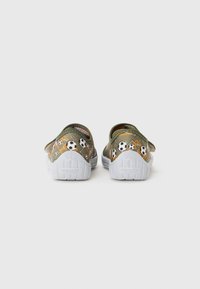 Chaussures en tissu vert avec semelles blanches, présentant des motifs de ballons de soccer et des accents orange sur les côtés. Design sans lacets. 