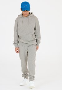 Grauer Sweatshirt mit Kapuze und Fronttasche, kombiniert mit passenden grauen Sweatpants. Weiße Sneaker und eine blaue Mütze mit Aufschrift vervollständigen das Outfit.