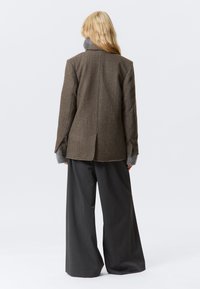 Bruine tweed blazer met een gestructureerd ontwerp, voorzien van een brede kraag en grijze laagjes manchetten, gecombineerd met losse grijze broek.