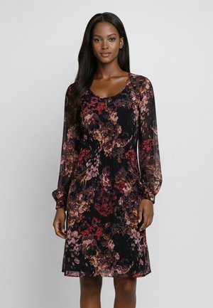 Femme portant une robe noire à manches longues et longueur genoux avec un motif floral multicolore, debout devant un fond blanc uni.